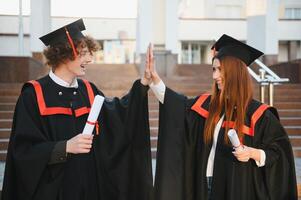 Portrait of happy graduates. Two friends in graduation caps and gowns standing outside university building with other students in background, holding diploma scrolls, and smiling photo