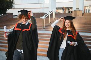 Portrait of happy graduates. Two friends in graduation caps and gowns standing outside university building with other students in background, holding diploma scrolls, and smiling photo