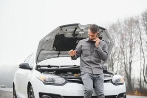 Man use a cellphone call garage in front of the open hood of a broken car on the road in the forest. Car breakdown concept. photo