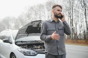 Sad driver calling car service, opening hood, having engine problem standing near broken car on the road. Car breakdown concept photo