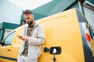 bearded caucasian man standing near an electric car that is charging and making time adjustments on a smartphone. photo