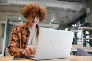 Portrait of Caucasian male freelancer in trendy apparel sitting at cafeteria table and doing remote work for programming design of public website, skilled software developer posing in coworking space. photo