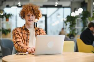 Portrait of Caucasian male freelancer in trendy apparel sitting at cafeteria table and doing remote work for programming design of public website, skilled software developer posing in coworking space. photo