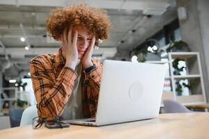 Portrait of Caucasian male freelancer in trendy apparel sitting at cafeteria table and doing remote work for programming design of public website, skilled software developer posing in coworking space. photo