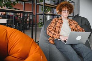 Portrait of Caucasian male freelancer in trendy apparel sitting at cafeteria table and doing remote work for programming design of public website, skilled software developer posing in coworking space. photo