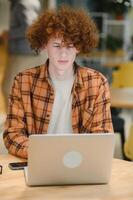 Portrait of Caucasian male freelancer in trendy apparel sitting at cafeteria table and doing remote work for programming design of public website, skilled software developer posing in coworking space. photo