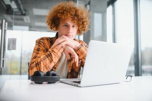 Portrait of Caucasian male freelancer in trendy apparel sitting at cafeteria table and doing remote work for programming design of public website, skilled software developer posing in coworking space. photo