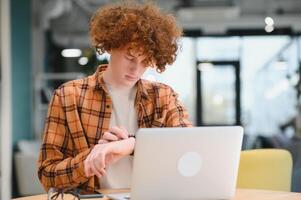 Portrait of Caucasian male freelancer in trendy apparel sitting at cafeteria table and doing remote work for programming design of public website, skilled software developer posing in coworking space. photo