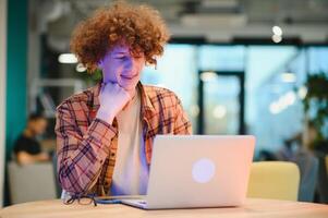 Portrait of Caucasian male freelancer in trendy apparel sitting at cafeteria table and doing remote work for programming design of public website, skilled software developer posing in coworking space. photo