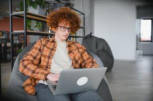 Portrait of Caucasian male freelancer in trendy apparel sitting at cafeteria table and doing remote work for programming design of public website, skilled software developer posing in coworking space. photo