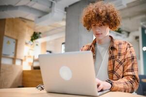 Portrait of Caucasian male freelancer in trendy apparel sitting at cafeteria table and doing remote work for programming design of public website, skilled software developer posing in coworking space. photo