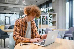 Portrait of Caucasian male freelancer in trendy apparel sitting at cafeteria table and doing remote work for programming design of public website, skilled software developer posing in coworking space. photo