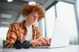 Portrait of Caucasian male freelancer in trendy apparel sitting at cafeteria table and doing remote work for programming design of public website, skilled software developer posing in coworking space. photo
