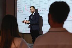 Business Conference Meeting Presentation. Businessman does Financial Analysis talks to Group of Businessspeople. Projector Screen Shows Stock Market Data, Investment Strategy, Revenue Growth photo