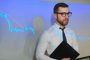 Stock Market Top Trader Looks at Projected Ticker Numbers and Graphs Running, Analysing Data to Make Best Sell. Behind Him Room Full of Screens and Statistics. photo