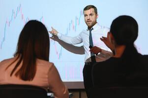Business Conference Meeting Presentation. Businessman does Financial Analysis talks to Group of Businessspeople. Projector Screen Shows Stock Market Data, Investment Strategy, Revenue Growth photo