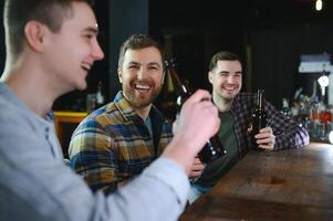 Three young men in casual clothes are smiling, holding bottles of beer while standing near bar counter in pub photo