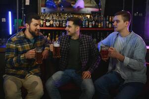 Three young men in casual clothes are smiling, holding bottles of beer while standing near bar counter in pub photo