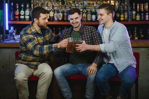 Three young men in casual clothes are smiling, holding bottles of beer while standing near bar counter in pub photo