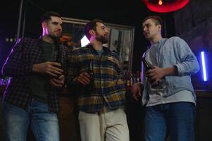 Three young men in casual clothes are smiling, holding bottles of beer while standing near bar counter in pub photo