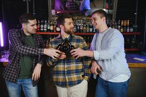 Three young men in casual clothes are smiling and clanging glasses of beer together while sitting at bar counter in pub photo