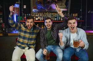 Soccer fans cheering for favourite team while enjoying drinks. Group of supporters celebrating victory while watching football match on tv in a club. Happy friends shouting while cheering for a goal. photo