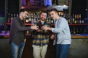 Three young men in casual clothes are smiling and clanging glasses of beer together while sitting at bar counter in pub photo