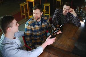 Three young men in casual clothes are smiling, holding bottles of beer while standing near bar counter in pub photo
