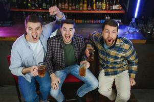 Soccer fans cheering for favourite team while enjoying drinks. Group of supporters celebrating victory while watching football match on tv in a club. Happy friends shouting while cheering for a goal. photo
