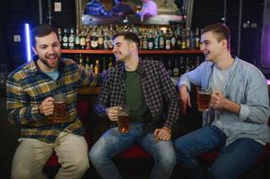 Three young men in casual clothes are smiling, holding bottles of beer while standing near bar counter in pub photo