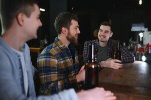 Three young men in casual clothes are smiling, holding bottles of beer while standing near bar counter in pub photo