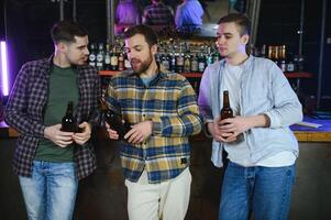 Three young men in casual clothes are smiling and clanging glasses of beer together while sitting at bar counter in pub photo