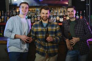 Three young men in casual clothes are smiling, holding bottles of beer while standing near bar counter in pub photo
