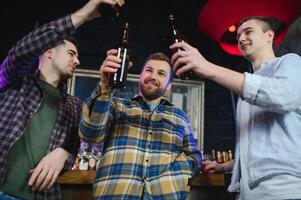 Three young men in casual clothes are smiling, holding bottles of beer while standing near bar counter in pub photo
