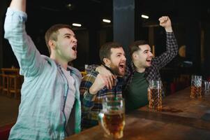 Soccer fans cheering for favourite team while enjoying drinks. Group of supporters celebrating victory while watching football match on tv in a club. Happy friends shouting while cheering for a goal. photo