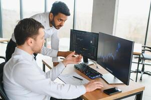 Two successful trader sitting in office and looking at screen, checking cryptocurrency information data on finance market graph, pointing on monitor photo