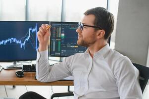 Man trader in formalwear sitting at desk in frot of monitors with charts and data at office browsing laptop checking documents analyzing stocks price changes concentrated. photo
