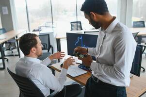 Two men traders sitting at desk at office together monitoring stocks data candle charts on screen analyzing price flow smiling cheerful having profit teamwork concept photo