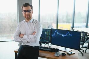 Man trader in formalwear sitting at desk in frot of monitors with charts and data at office browsing laptop checking documents analyzing stocks price changes concentrated. photo