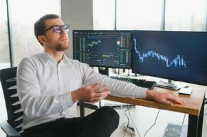Man trader in formalwear sitting at desk in frot of monitors with charts and data at office browsing laptop checking documents analyzing stocks price changes concentrated. photo
