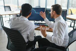 Two men traders sitting at desk at office together monitoring stocks data candle charts on screen analyzing price flow smiling cheerful having profit teamwork concept photo