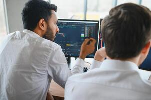 Two successful trader sitting in office and looking at screen, checking cryptocurrency information data on finance market graph, pointing on monitor photo