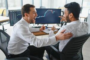 Two men traders sitting at desk at office together monitoring stocks data candle charts on screen analyzing price flow smiling cheerful having profit teamwork concept photo