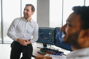 Two men traders sitting at desk at office together monitoring stocks data candle charts on screen analyzing price flow smiling cheerful having profit teamwork concept photo