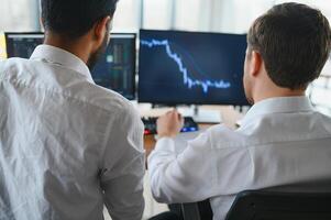 Two successful trader sitting in office and looking at screen, checking cryptocurrency information data on finance market graph, pointing on monitor photo