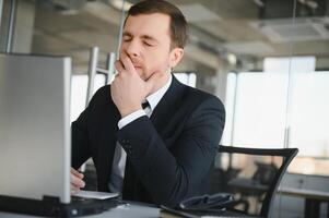 Young stressed handsome businessman working at desk in modern office shouting at laptop screen and being angry about financial situation, jealous of rival capabilities, unable to meet client needs photo