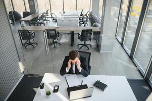 Young stressed handsome businessman working at desk in modern office shouting at laptop screen and being angry about financial situation, jealous of rival capabilities, unable to meet client needs photo