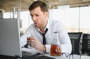 Young stressed handsome businessman working at desk in modern office shouting at laptop screen and being angry about financial situation, jealous of rival capabilities, unable to meet client needs photo