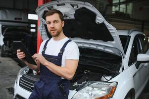Bearded mechanic in overalls standing in garage of a car salon and holding tablet. He is about to diagnostic breakdown. photo