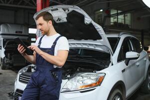 Bearded mechanic in overalls standing in garage of a car salon and holding tablet. He is about to diagnostic breakdown. photo